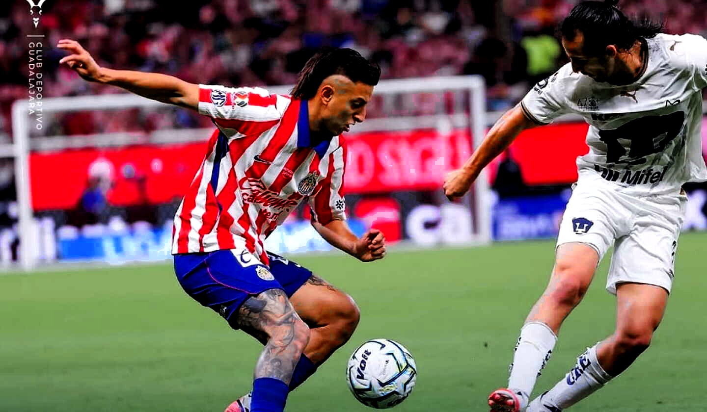 Jugador de Chivas, rayas rojas y azules, disputa el balón con uno de Pumas de blanco; césped y portería a color