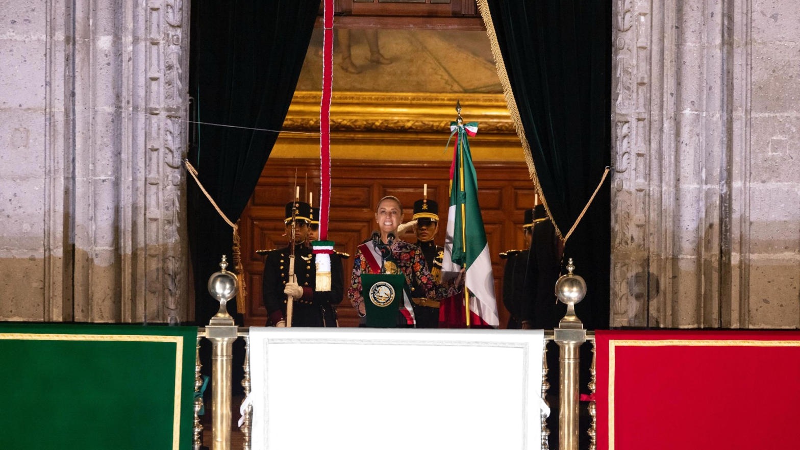 Claudia Sheinbaum, en escala de grises, con banda presidencial en el balcón de Palacio Nacional; banderas, recinto y escolta a color